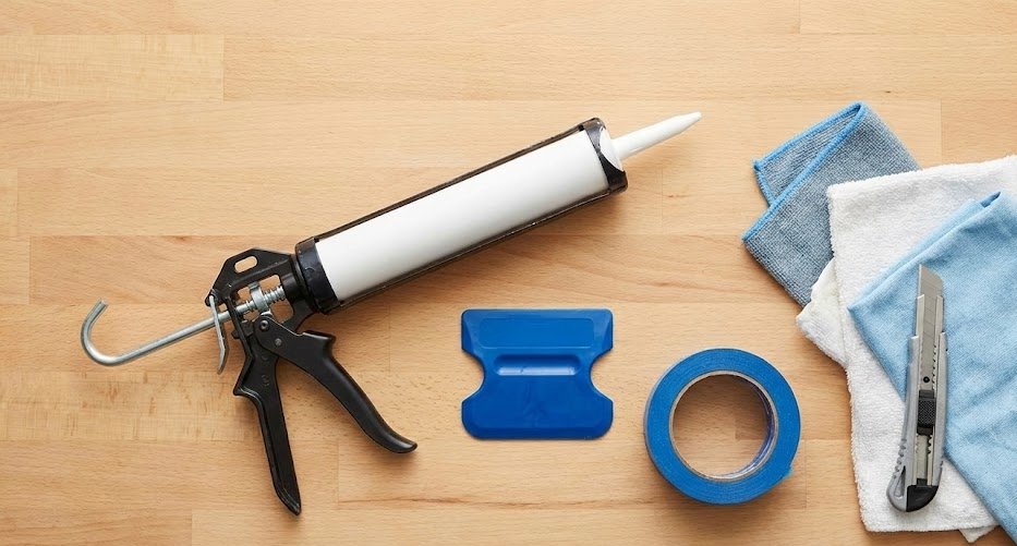 Hand using a caulk smoothing tool to create a clean, even bead of caulk along a bathtub edge with blue painter's tape